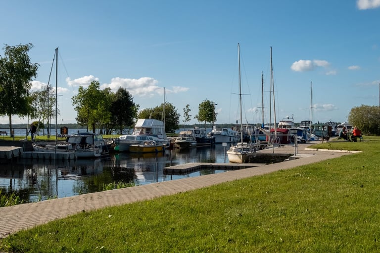 Marina with sailboats docked along a waterway, green grass in foreground, trees and blue sky above
